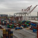 Overhead shot of a bustling container port with cranes and stacked shipping containers under a cloudy sky.